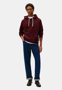 Felpa con cappuccio color bordeaux e cordini bianchi, abbinata a jeans blu. Il modello sta in piedi, mostrando un look casual a strati e scarpe nere.