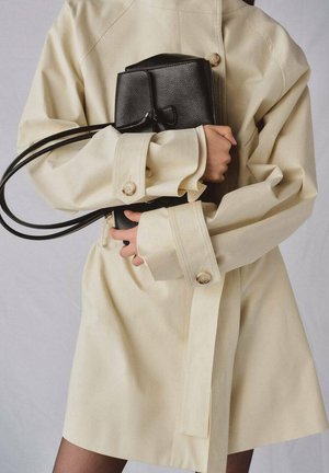 Person wearing beige trench coat holding a black textured leather handbag with double straps against a light background.