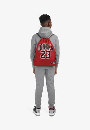 Sac à dos à cordon rouge avec "JORDAN" et "23" imprimés en noir, porté sur un sweat-shirt gris et un pantalon de survêtement. Des chaussures noires et rouges complètent la tenue.