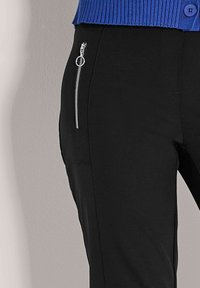 Zwarte broek met een verticale zilveren ritszak aan de zijkant, gedragen met een blauw ribbelgebreid topje met een zichtbare knop.