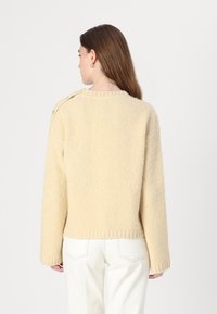 Crème gebreide trui met een gestructureerde afwerking, voorzien van een ronde halslijn en knoopdetail op de schouder. G gedragen met witte broek.