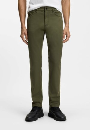 Mężczyzna w dopasowanych oliwkowozielonych spodniach i czarnych sneakersach, stojący na jasnym, jednolitym tle.