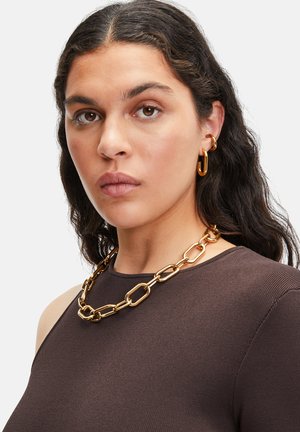 Collier en chaîne dorée avec des maillons allongés, porté sur un haut en ribbed brun, complété par des boucles d'oreilles en or en forme de hoop. Design simple et moderne.