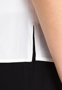 Blusa blanca texturizada con dobladillo dividido, combinada con tela negra. Piel visible en el brazo donde la blusa se une con la manga.