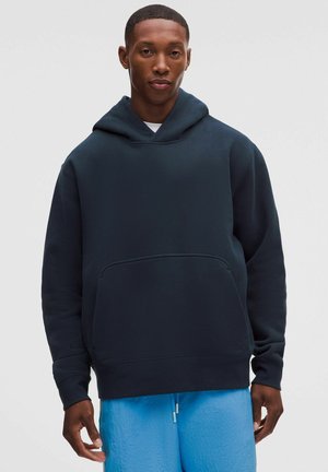 Hombre con sudadera con capucha azul marino oscuro y pantalones deportivos azul brillante de pie frente a un fondo claro liso.