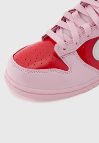 Ροζ αθλητικό παπούτσι με κόκκινο γυαλιστερό κάλυμμα στα δάχτυλα, οπές και ανοιχτό ροζ λεπτομέρειες. Διαθέτει επίπεδα κορδόνια και στρογγυλεμένη σόλα για υποστήριξη.