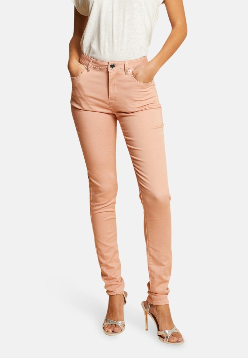 Jean slim rose avec fermeture à boutons, cinq poches et une texture lisse, associé à des talons hauts argentés à brides.