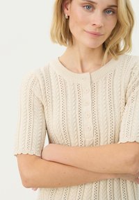 Haut en tricot beige à manches courtes, présentant un design en relief de câbles, cinq boutons sur le devant et un col rond avec des bords festonnés.