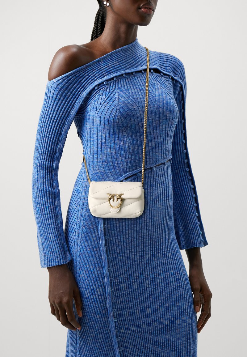 Borsa a tracolla bianca trapuntata con accessori dorati, caratterizzata da un emblema a testa di cavallo. Indossata con un vestito blu strutturato con dettagli a coste.