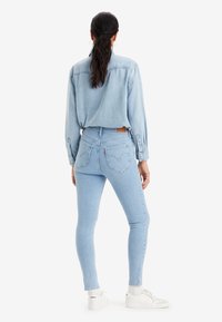 Pantalones vaqueros ajustados de mezclilla azul claro con cintura alta, que presentan bolsillos traseros y una etiqueta marrón en la cintura, combinados con una camisa de color azul claro a juego.