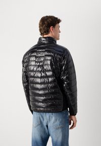 Man in een glanzende zwarte gewatteerde jas, met zijn rug naar ons gekeerd, in combinatie met lichtblauwe spijkerbroek, tegen een eenvoudige lichte achtergrond.
