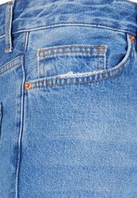 Los jeans de mezclilla azul claro presentan un corte clásico con un bolsillo trasero, detalles de costura y una textura ligeramente desvaída. Remaches de cobre adornan los bolsillos.