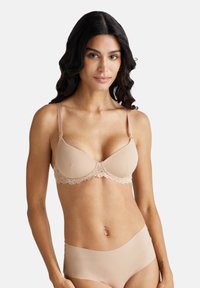 Sostegno in pizzo beige, coppe sagomate, spalline regolabili e una consistenza liscia. Indossato con biancheria intima beige coordinata.