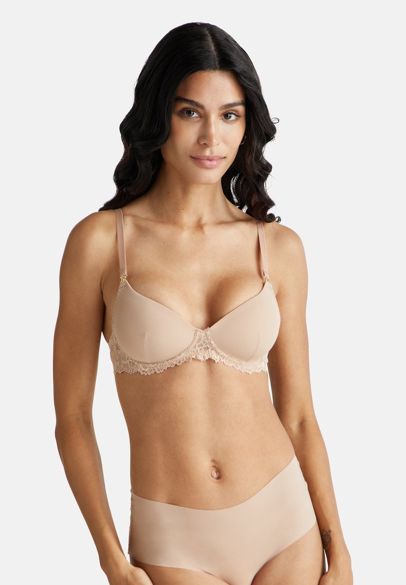 Sostegno in pizzo beige, coppe sagomate, spalline regolabili e una consistenza liscia. Indossato con biancheria intima beige coordinata.