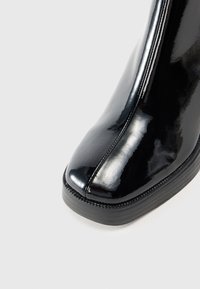 Gros plan d’une botte en cuir noir brillant à bout carré, avec des coutures visibles et une semelle épaisse en caoutchouc noir sur un fond clair.