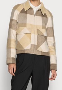 Ruskean ja beigen ruudullinen villatakki, jossa on kaulus, vetoketjusuljenta ja kaksi etutaskua; teksturoitu kangas kontrastoivilla kuoseilla.