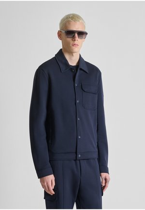 Blond man wearing black sunglasses, a dark navy button-up jacket with chest pocket, and matching pants against a plain gray background.