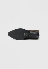 Barna velúr csizma kerek orral, lapos fekete gumitalppal és texturált részletekkel, a talpon kör alakú logóval.