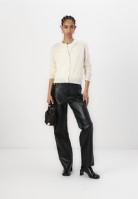 Gilet crème avec des boutons, associé à un pantalon en cuir noir taille haute. Porte un petit sac sombre ; des bottines noires à talons viennent compléter le look.