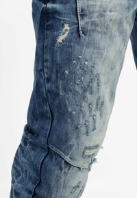 Faded blue denim jeans with distressed accents, frayed patches, and a tapered fit. Features a smooth texture and light wear evident.