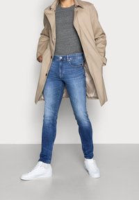 Beige trenchcoat över en grå skjorta, blå slim-fit jeans och vita sneakers. Kappan har en slät textur med knappdetaljer.