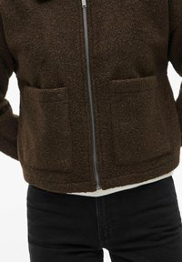 Veste en polaire marron avec une fermeture éclair à l'avant. Elle comporte deux grandes poches et une surface texturée. Portée par-dessus un haut blanc et un pantalon noir.