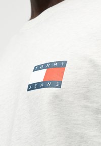 Ανοιχτό γκρι βαμβακερό μπλουζάκι με λογότυπο σε σχήμα πλακιδίου με κόκκινες, λευκές και μπλε ορθογώνιες και το κείμενο "TOMMY JEANS" σε έντονη γραμματοσειρά.