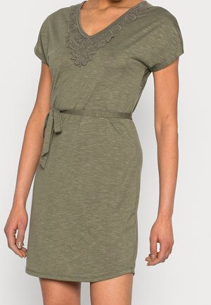 Frau trägt ein olivgrünes Kleid mit kurzen Ärmeln, Spitzen-V-Ausschnitt und einem Stoffgürtel, der in der Taille gebunden ist, steht vor schlichtem Hintergrund.