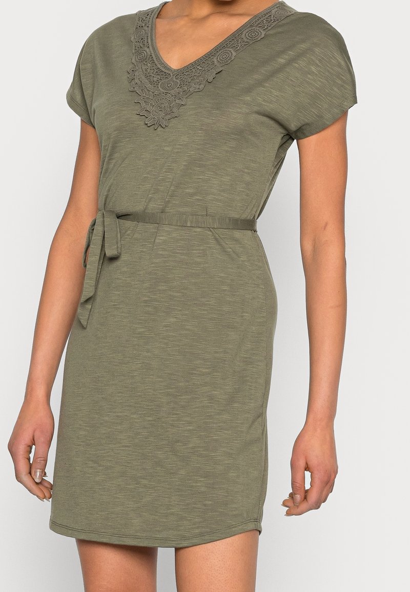 Femme portant une robe vert olive à manches courtes avec un décolleté en V en dentelle et une ceinture en tissu nouée à la taille, debout devant un fond uni.