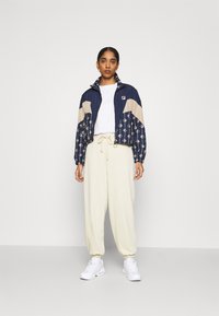 Giacca zip-up navy e beige con motivi geometrici, abbinata a pantaloni jogger crema e sneaker bianche. Tessuto leggero e casual.