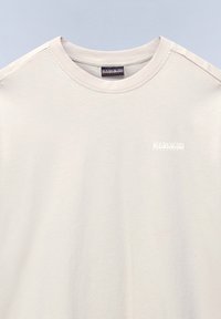 Lys beige bomulds t-shirt med rund halsudskæring og korte ærmer. Har et lille hvidt logo på brystområdet. Glat tekstur.