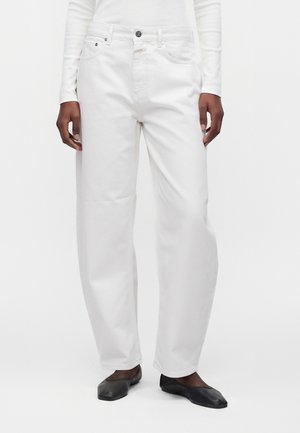 Person iført løse hvide jeans, hvid langærmet top og sorte flade sko står mod en ensfarvet hvid baggrund.