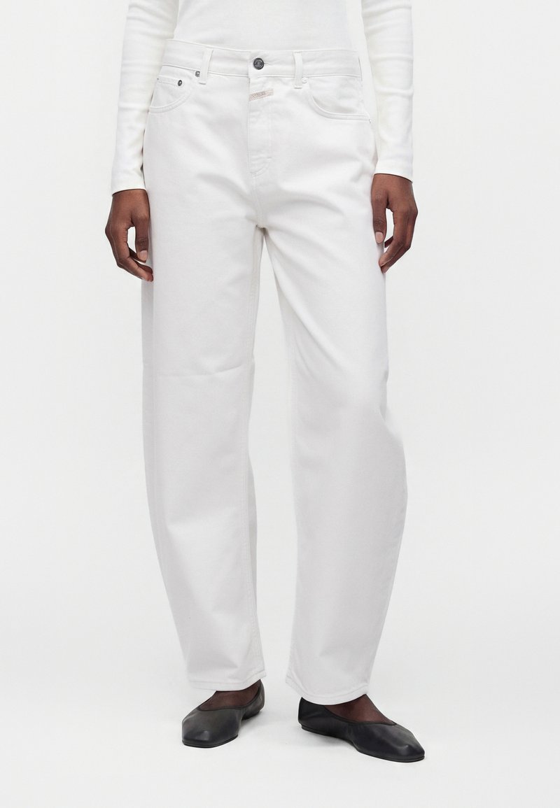 Personne portant un jean blanc ample, un haut blanc à manches longues et des chaussures plates noires, debout sur un fond blanc uni.