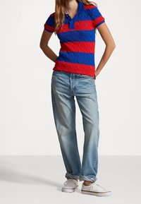 Polo tričko s červenými a modrými pruhy, se vzorem jako pletenina, v kombinaci se světle modrými džíny a bílými teniskami. Krátké rukávy a detail límce jsou viditelné.