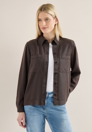 Blonde Frau, die eine dunkelbraune Jacke mit Knöpfen über einem weißen Hemd trägt, kombiniert mit hellblauen Jeans, stehend vor einem neutralen Hintergrund.