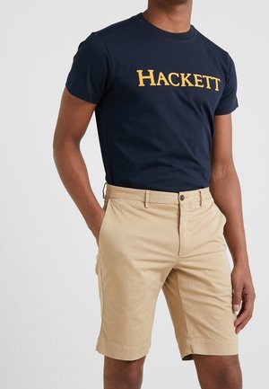 Marineblaues Baumwoll-T-Shirt mit goldgesticktem "HACKETT"-Logo, kombiniert mit beigen maßgeschneiderten Shorts, die über Fronttaschen und eine glatte Textur verfügen.