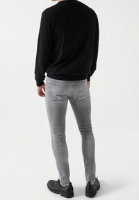 Personne portant un haut à manches longues noir, un jean slim gris clair et des chaussures noires à lacets, debout devant un fond blanc uni.