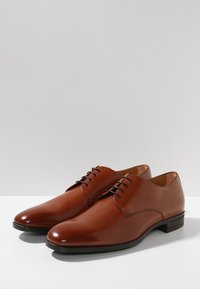 Chaussures de ville pour hommes en cuir marron avec des lacets noirs,  bout rond et semelles basses, placées côte à côte sur une surface blanche.
