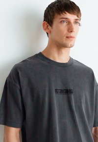 Camiseta de algodón de gran tamaño en gris oscuro con mangas cortas y escote redondo. Presenta un sutil logotipo gráfico negro en el pecho.