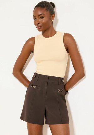 Kvinde med mørk hud og hår i knold iført beige ærmeløs top og mørkebrune shorts med høj talje med dekorative knapper og kæder.