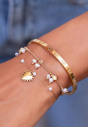 Quattro braccialetti a strati: un sottile bracciale in oro con design a cuore, una catena in oro con un ciondolo a cuore e perle, e una catena in oro con perline.