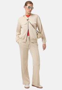 Béžová bomber bunda s dvěma předními kapsami, doplněná béžovými kalhotami. Pod ní je červená košile a malá kabelka s logovým popruhem.