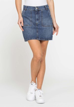 Persona che indossa una minigonna di jeans blu con tasche frontali, abbinata a sneakers bianche e una maglietta bianca infilata dentro.