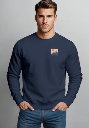 Mann mit kurzen braunen Haaren, der einen marineblauen Kapuzenpullover mit einer kleinen Grafik von Bergen und einer Sonne auf der Brust trägt, posiert mit den Händen in den Hosentaschen.
