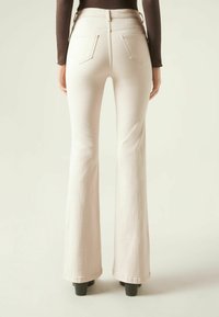 Pantalons flare beige clair avec une texture lisse, taille haute et deux poches arrière. Ourlet légèrement évasé sur des talons noirs.