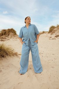 Femme portant une chemise en jean bleu clair et un pantalon à jambes larges, debout pieds nus sur une plage de sable avec de l'herbe de dune sous un ciel partiellement nuageux.