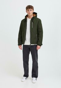 Giacca verde con zip e cappuccio, due tasche laterali e interno grigio. Indossata sopra una maglietta bianca, abbinata a jeans scuri e sneakers bianche.