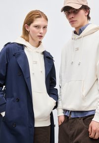 Νεαρή γυναίκα φορώντας ναυτικό μπλε καμπαρντίνα πάνω από κρεμ φούτερ και νεαρός άνδρας σε κρεμ φούτερ, μπλε καρό πουκάμισο, καφέ παντελόνι και μπεζ καπέλο στέκονται δίπλα-δίπλα.