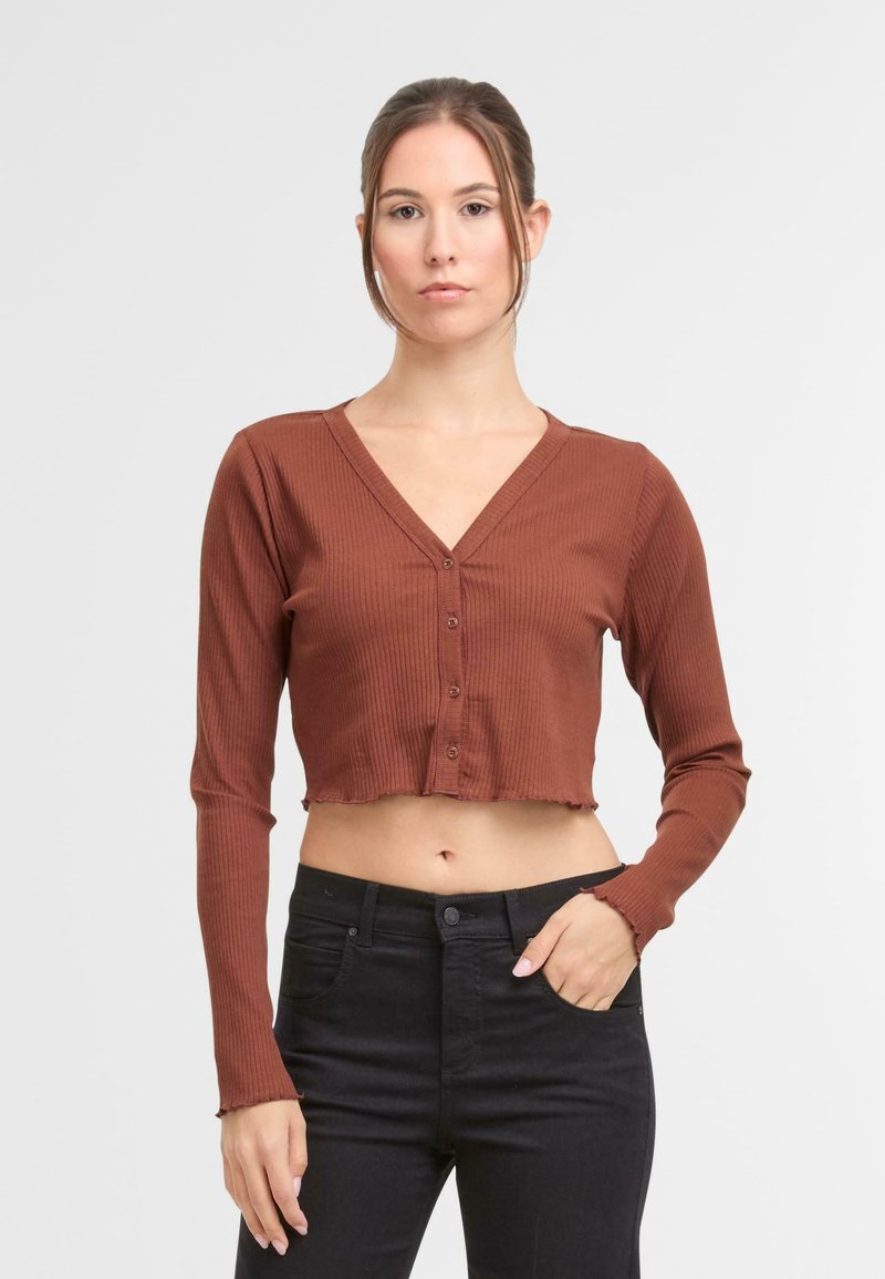 Smeđi rebrasti crop top s dugim rukavima i V-izrezom, s gumbima sprijeda i sirovim rubom. Uparen s crnim trapericama visokog struka.