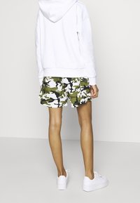 Felpa bianca con cappuccio, pantaloni corti mimetici verdi e bianchi, e sneakers bianche. I dettagli notevoli includono un fit rilassato e una tasca visibile sui pantaloni.